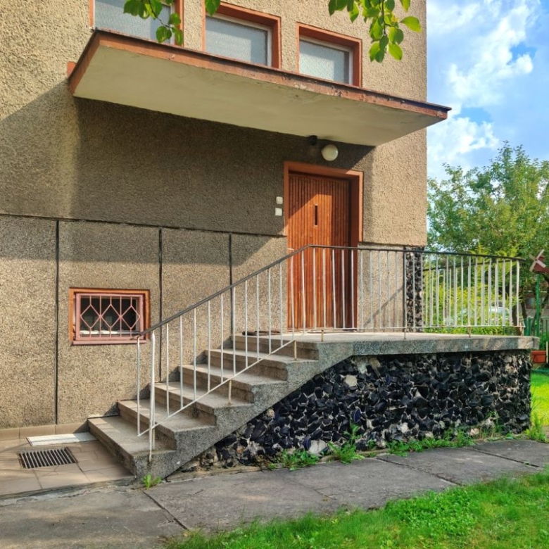 Entrance to a beige apartment building with a wooden door reached by concrete stairs and a metal handrail; stonework along the base and a small barred window on the wall.