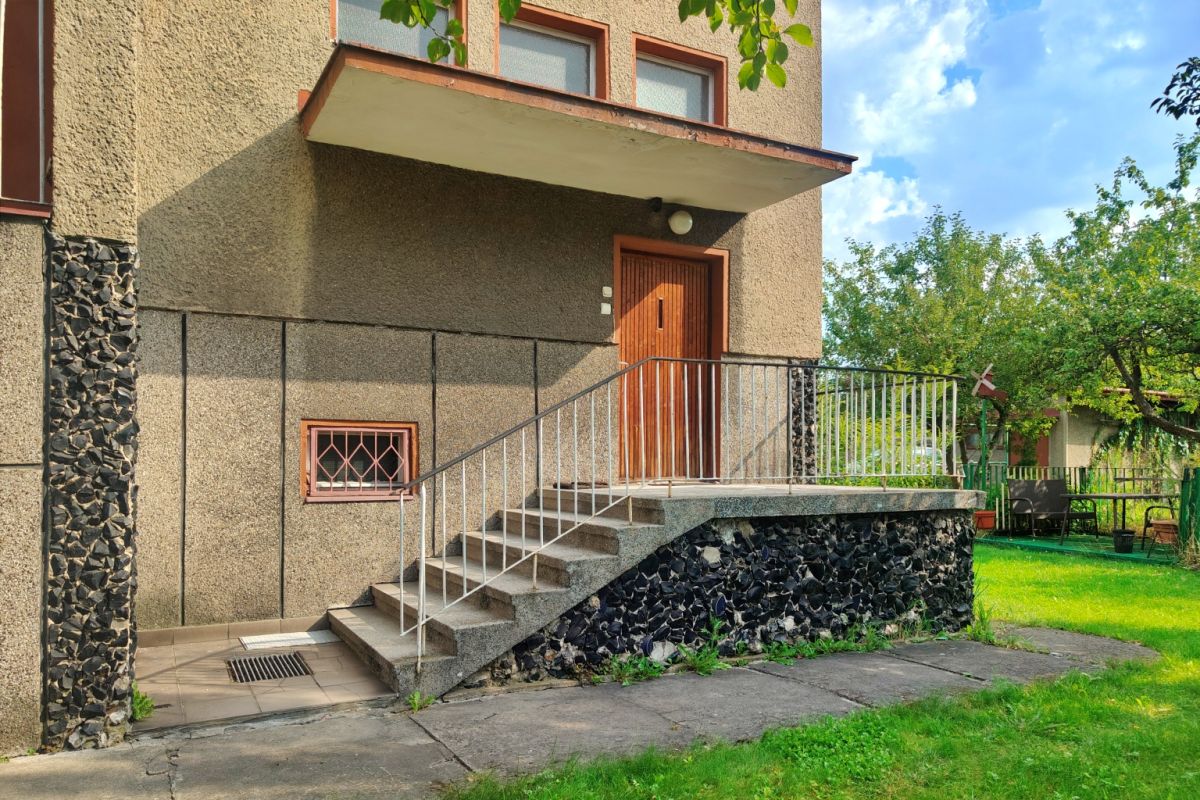 Entrance to a beige apartment building with a wooden door reached by concrete stairs and a metal handrail; stonework along the base and a small barred window on the wall.