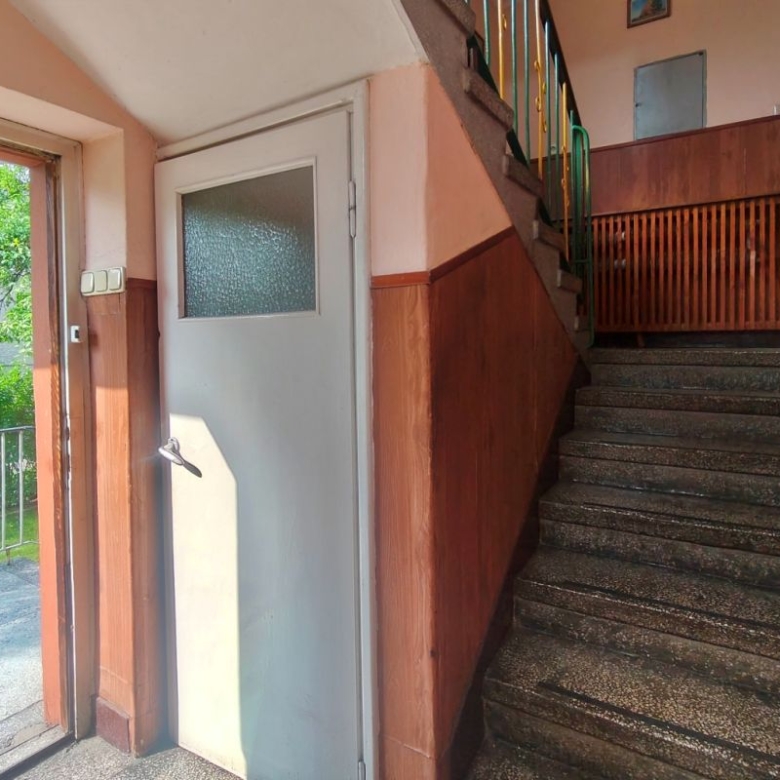 Interior stairwell with brown wood paneling, terrazzo stairs, and a gray exterior door; open door shows a green outdoor area with trees and railing outside.