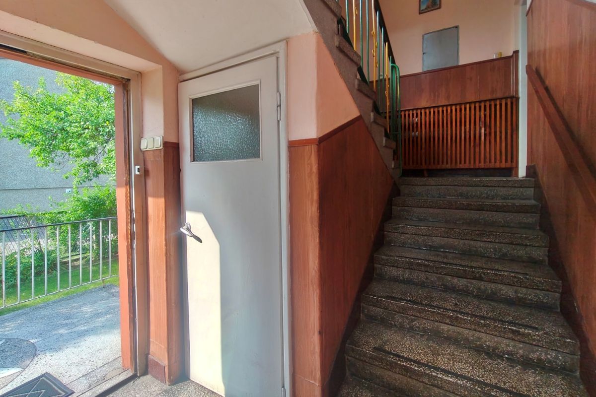 Interior stairwell with brown wood paneling, terrazzo stairs, and a gray exterior door; open door shows a green outdoor area with trees and railing outside.