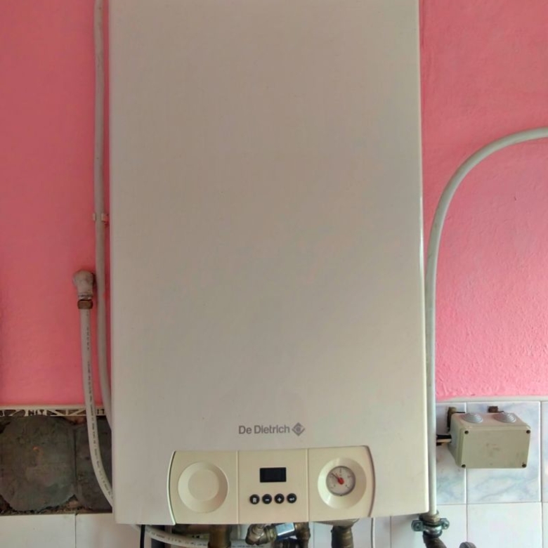 White wall-mounted boiler (De Dietrich) with exposed pipes and a pressure gauge below, mounted on a pink wall in a utility area.
