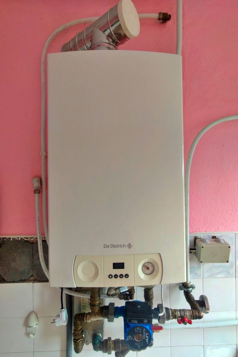White wall-mounted boiler (De Dietrich) with exposed pipes and a pressure gauge below, mounted on a pink wall in a utility area.