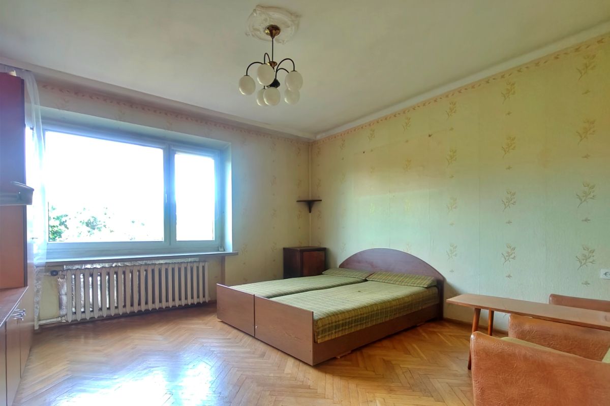 Sunlit bedroom with a wooden double bed at center, green plaid bedding, and light-colored walls with subtle floral wallpaper.