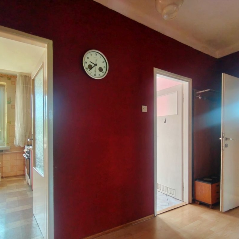 Dark burgundy hallway with a round clock, open doors lead to a sunlit kitchen on the left and another room on the right paneling.
