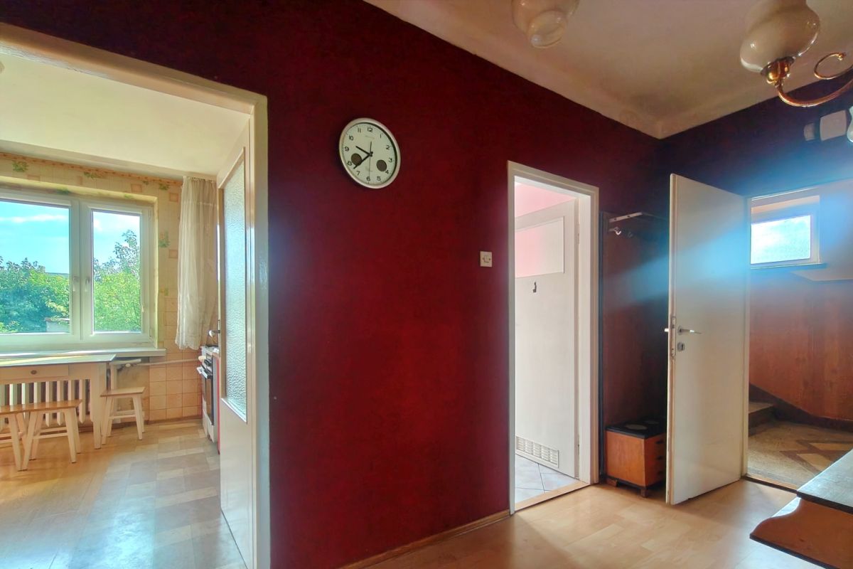 Dark burgundy hallway with a round clock, open doors lead to a sunlit kitchen on the left and another room on the right paneling.