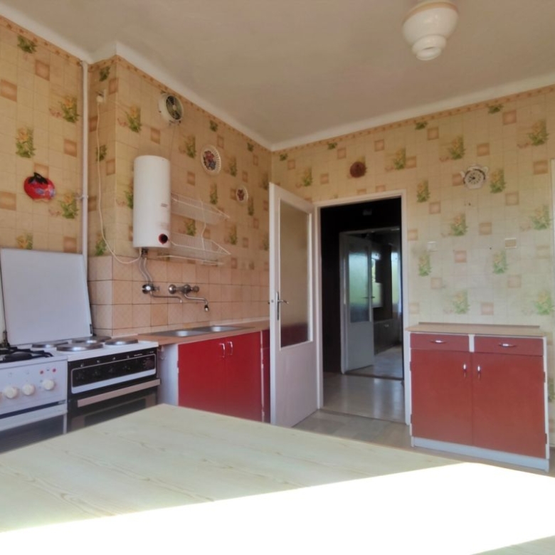 Old-fashioned kitchen with a gas range, red lower cabinets, and a light wooden table in the foreground; beige patterned wallpaper covers the walls all around.