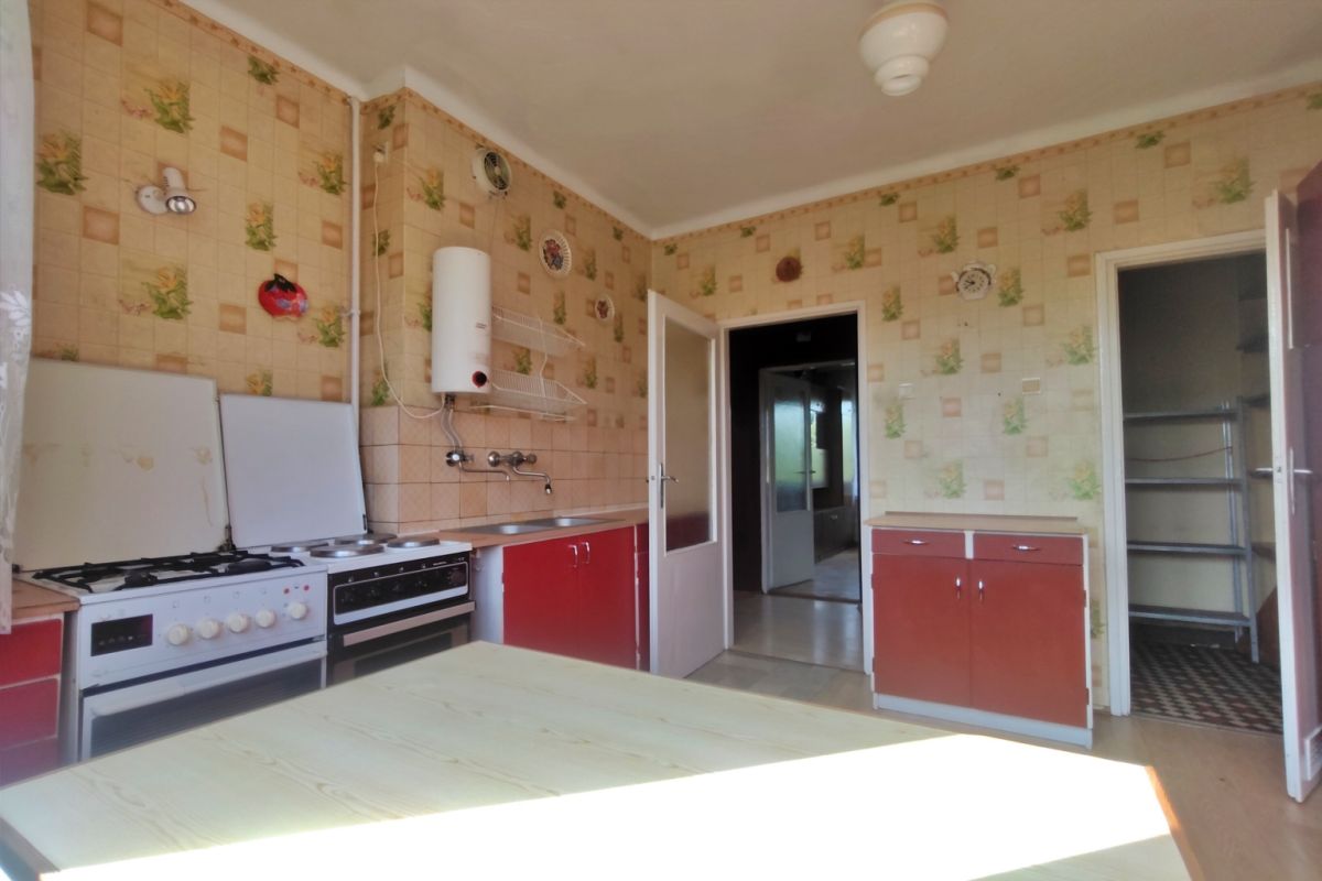 Old-fashioned kitchen with a gas range, red lower cabinets, and a light wooden table in the foreground; beige patterned wallpaper covers the walls all around.
