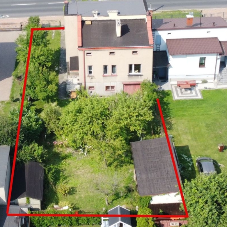 Aerial view of a rectangular residential property outlined in red, showing trees, a lawn, and two sheds between neighboring houses on a street edge.