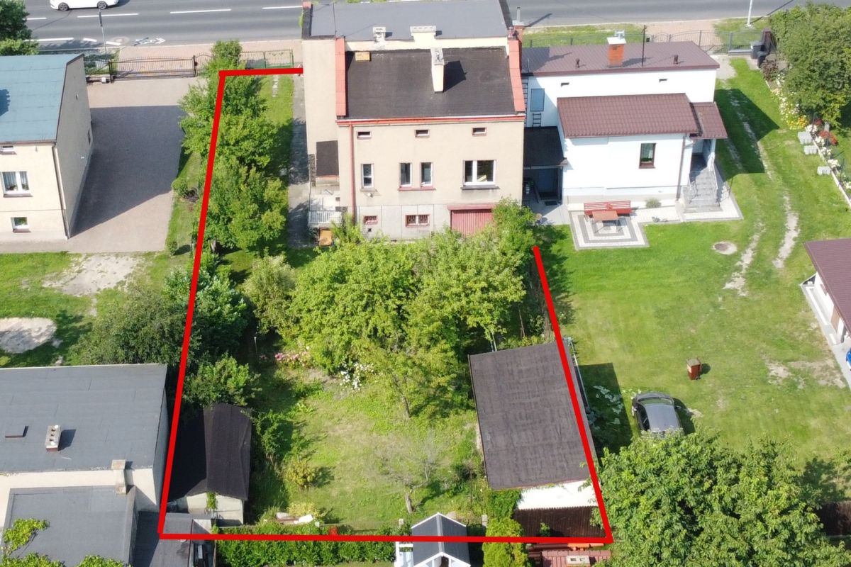Aerial view of a rectangular residential property outlined in red, showing trees, a lawn, and two sheds between neighboring houses on a street edge.