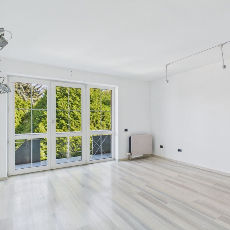 Bright empty room with light wood floors and white walls, large glass doors to a balcony and greenery outside.