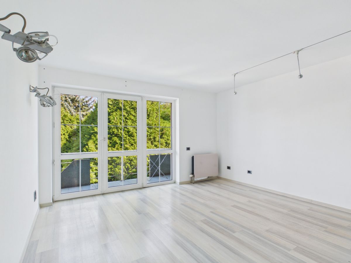 Bright empty room with light wood floors and white walls, large glass doors to a balcony and greenery outside.