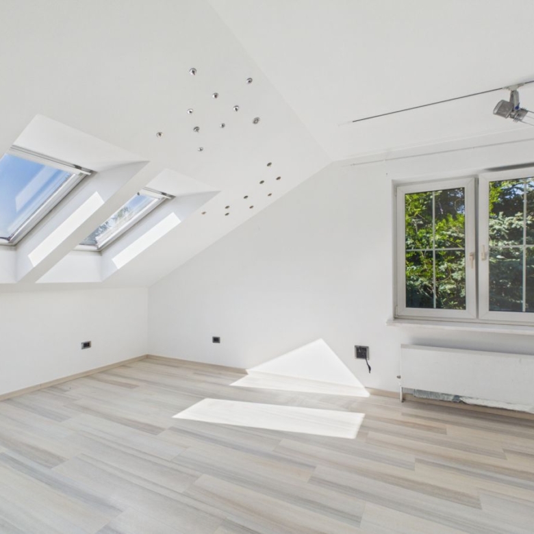 Bright empty attic room with slanted white walls, three skylights on the left, a large window on the right, and light wood flooring.