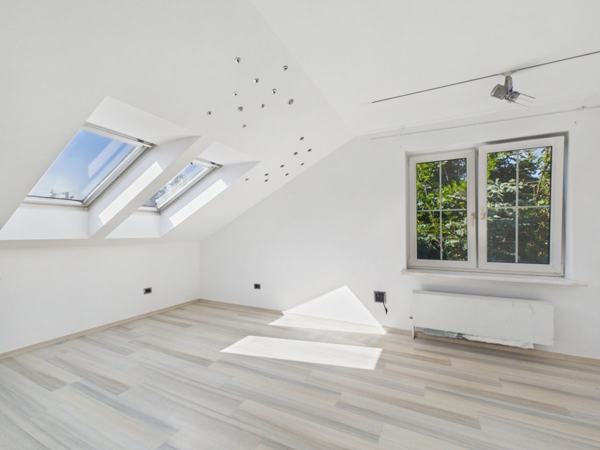 Bright empty attic room with slanted white walls, three skylights on the left, a large window on the right, and light wood flooring.