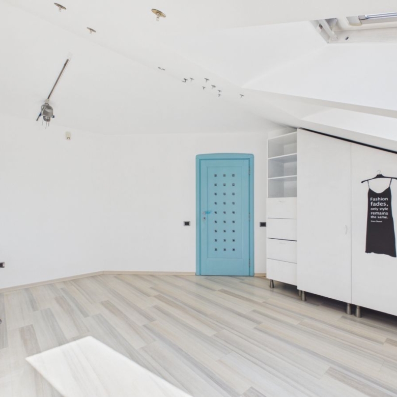 Bright white attic studio with light wood floor, blue door, white cabinets, and a black dress poster on the wardrobe.