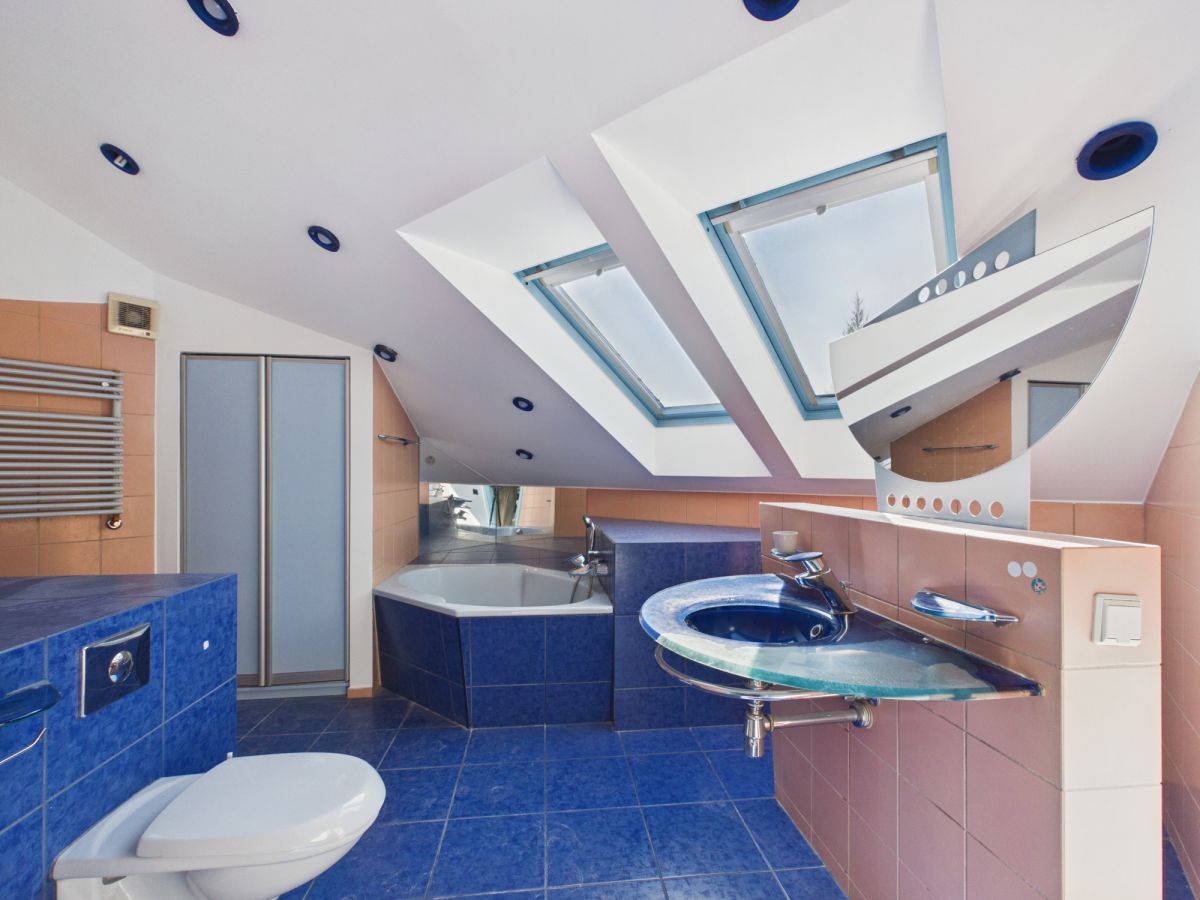 Bright modern bathroom with blue tiled floor and walls, a corner bathtub, and a blue glass sink under angled skylights in a white ceiling.
