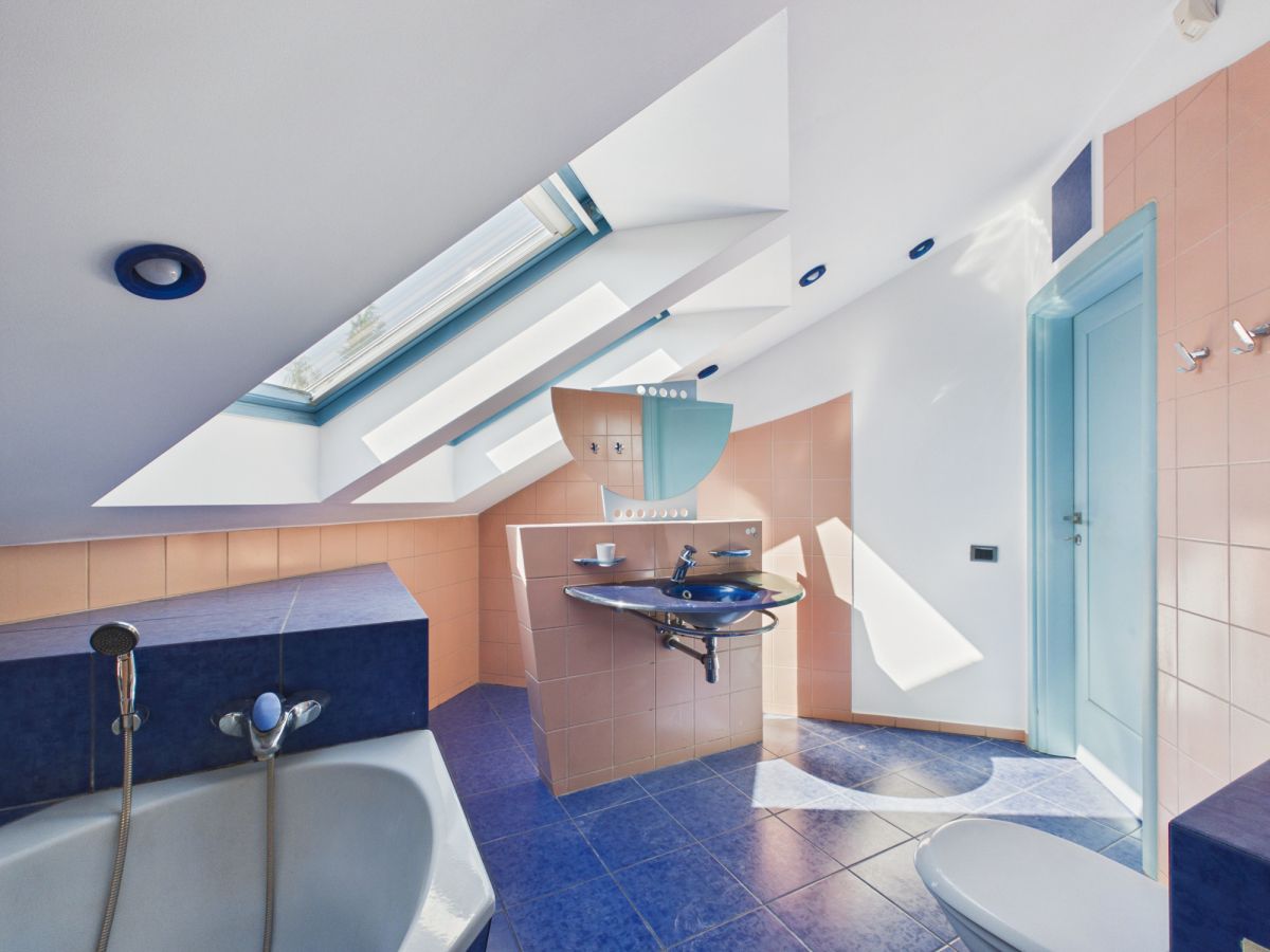 Bright modern bathroom with peach tile walls, blue accents, and a skylight casting geometric light on a pedestal sink area and bathtub.