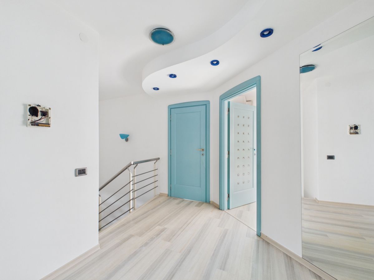 Bright, modern hallway with light wood floors, white walls, and turquoise door frames; curved ceiling with blue recessed lights.