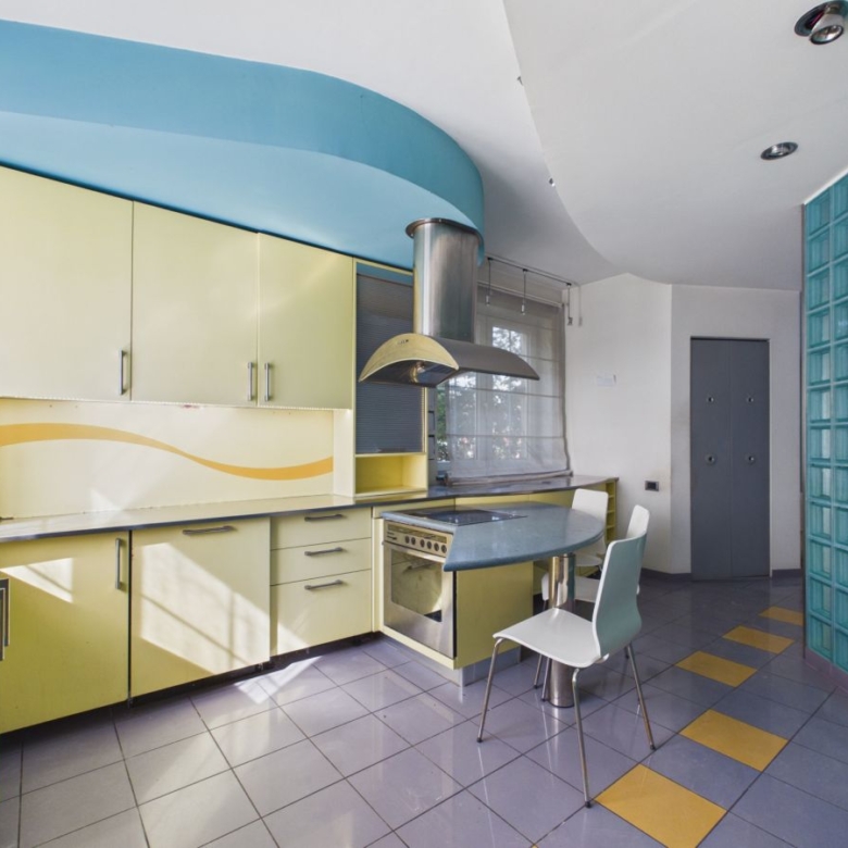 Retro-style kitchen with yellow cabinets, blue curved ceiling, and a blue countertop island with a stove. Glass block wall on the right.