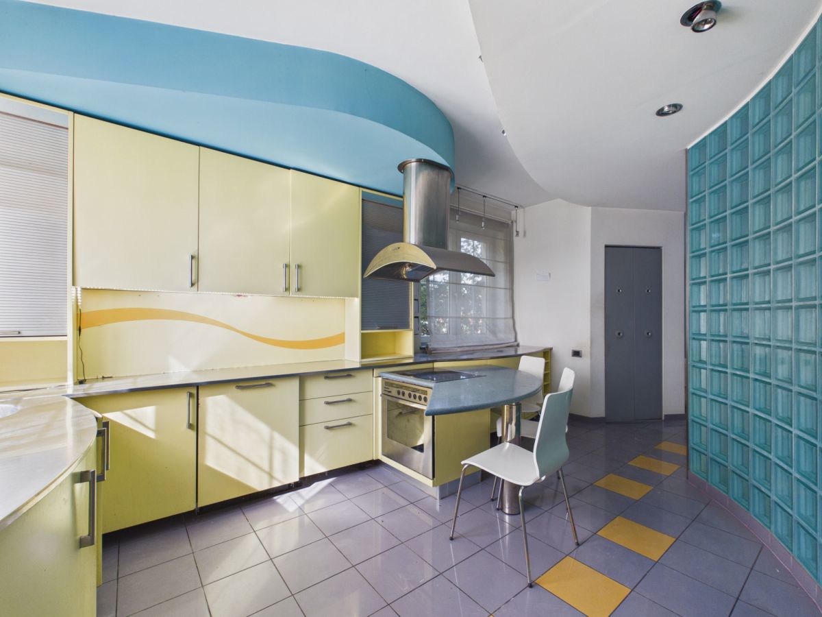 Retro-style kitchen with yellow cabinets, blue curved ceiling, and a blue countertop island with a stove. Glass block wall on the right.