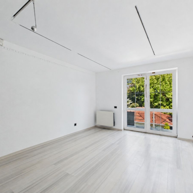 Bright, empty room with white walls and light wood floor; large glass doors open to a balcony with green trees outside.