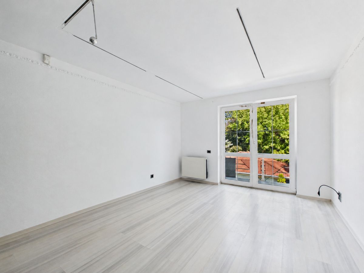 Bright, empty room with white walls and light wood floor; large glass doors open to a balcony with green trees outside.