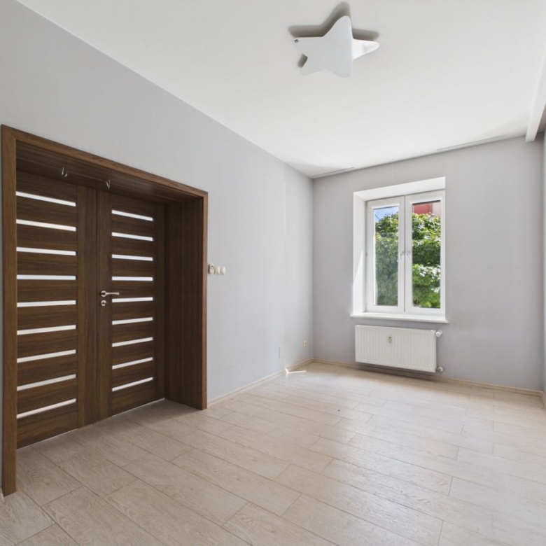 Empty light-filled room with a large wooden double door, gray walls, and a window with a radiator beneath it; star-shaped ceiling light fixture present.