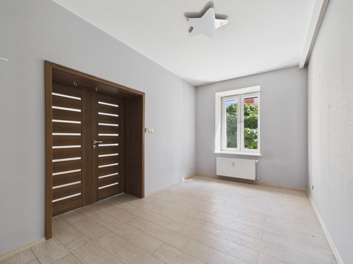 Empty light-filled room with a large wooden double door, gray walls, and a window with a radiator beneath it; star-shaped ceiling light fixture present.