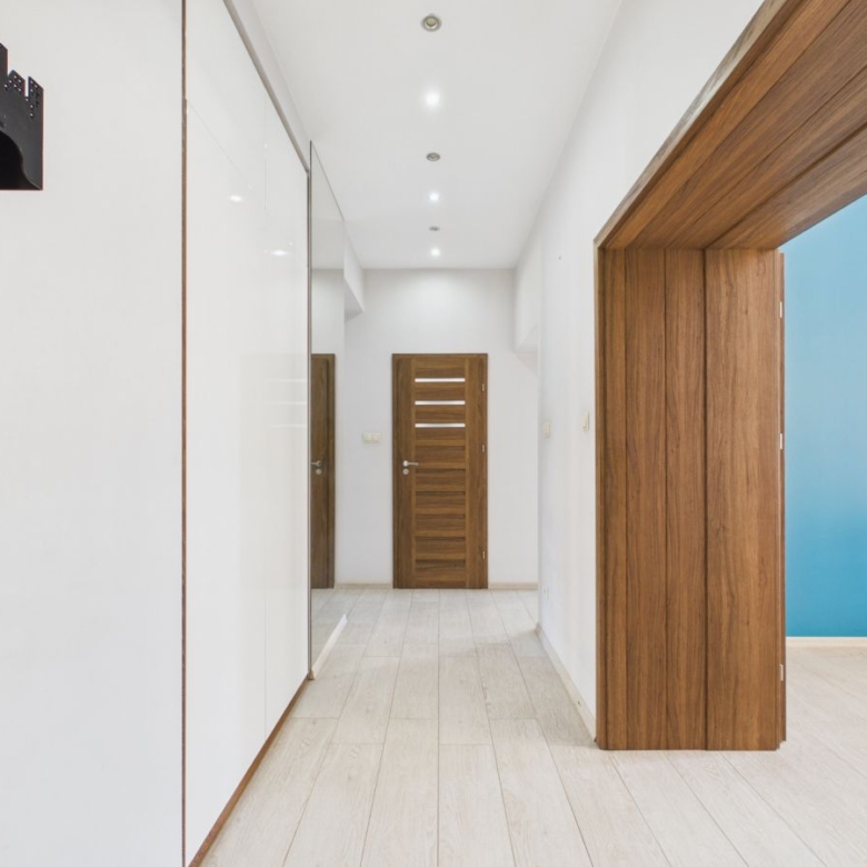 Bright hallway with white walls, light wood flooring, and recessed ceiling lights; a series of wooden doors lines the hall, with a blue-walled room visible through an open doorway on the right.