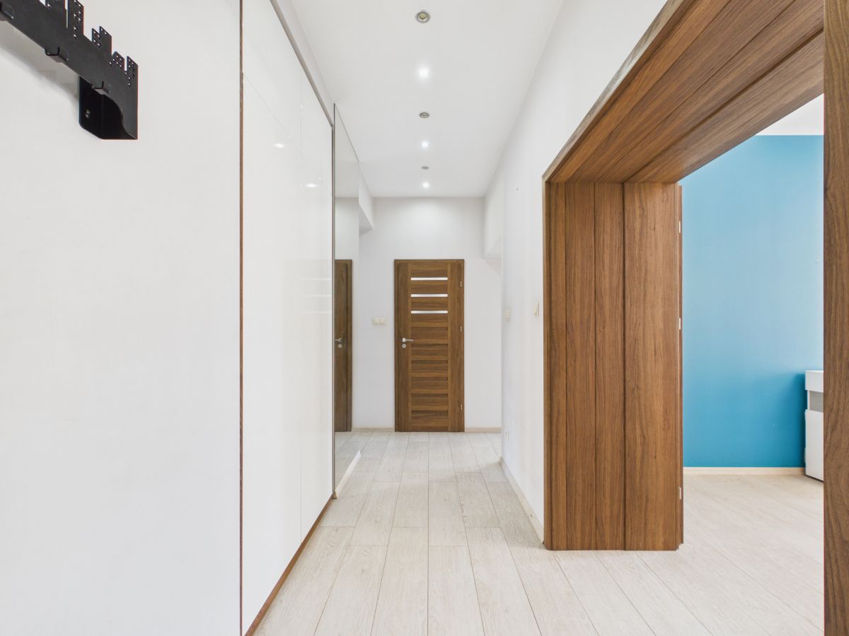 Bright hallway with white walls, light wood flooring, and recessed ceiling lights; a series of wooden doors lines the hall, with a blue-walled room visible through an open doorway on the right.