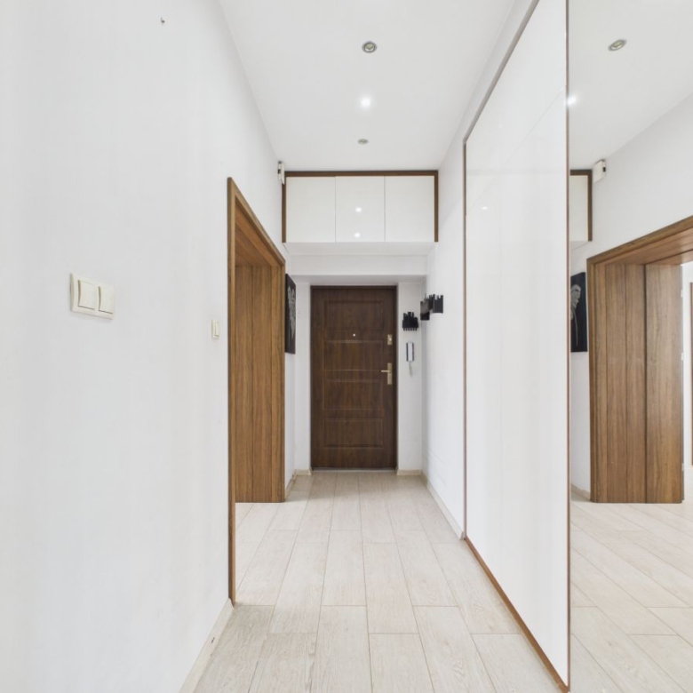 Bright minimalist hallway with light wood flooring, white walls, and a dark wooden front door at the end; recessed ceiling lights and built-in white cabinets above. A large mirrored wardrobe runs along the right wall, reflecting the scene.