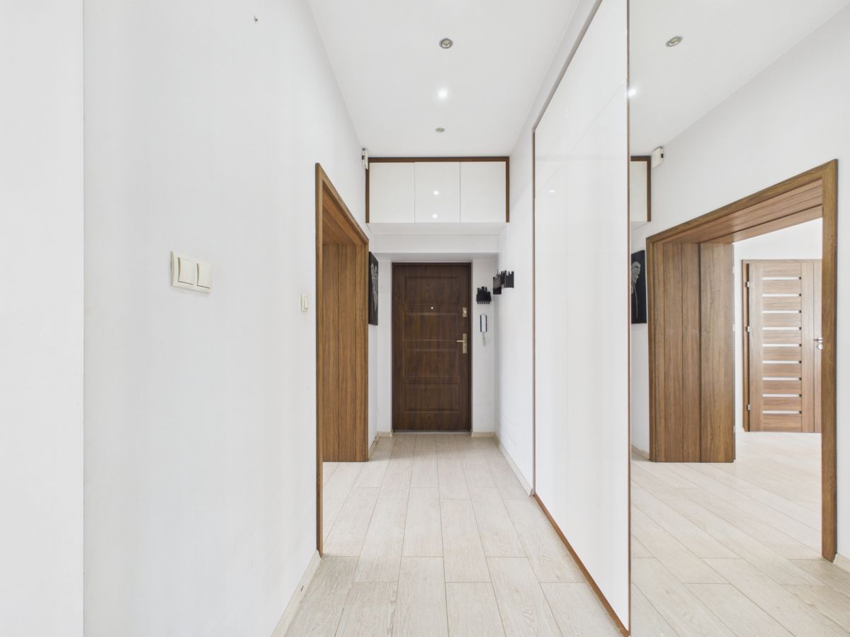 Bright minimalist hallway with light wood flooring, white walls, and a dark wooden front door at the end; recessed ceiling lights and built-in white cabinets above. A large mirrored wardrobe runs along the right wall, reflecting the scene.