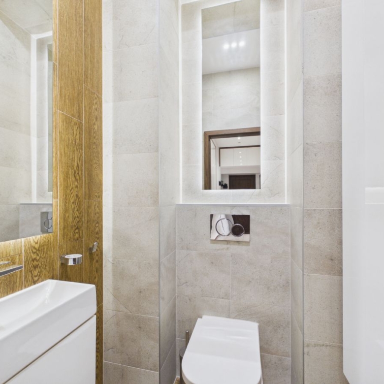 Modern bathroom with beige stone tiles, white wall-hung toilet, and rectangular sink beneath a large mirror with a wooden frame panel behind it.