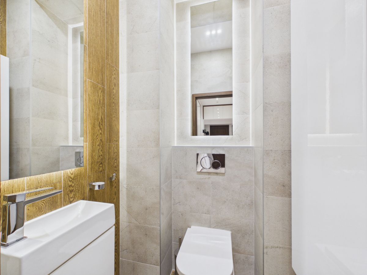 Modern bathroom with beige stone tiles, white wall-hung toilet, and rectangular sink beneath a large mirror with a wooden frame panel behind it.