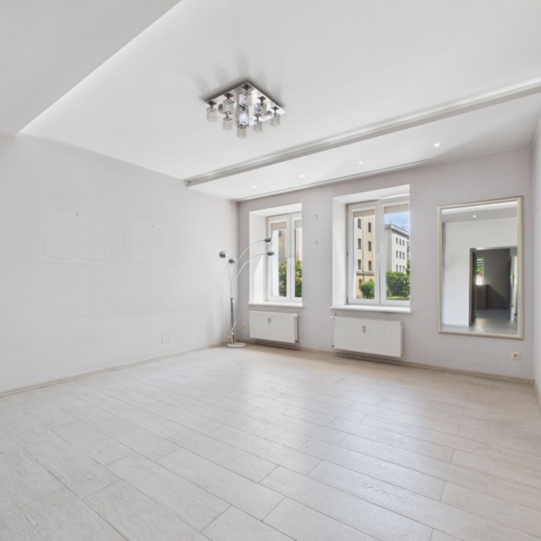 Bright, empty living room with light wood floors, white walls, and a stone accent wall on the right; three windows and a tall mirror reflect the space.