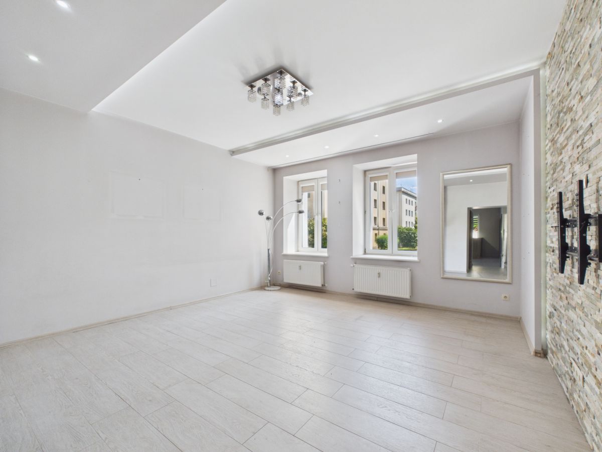 Bright, empty living room with light wood floors, white walls, and a stone accent wall on the right; three windows and a tall mirror reflect the space.