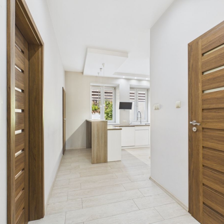 Bright hallway with light wood doors leading to a modern white kitchen with a window view outside far end