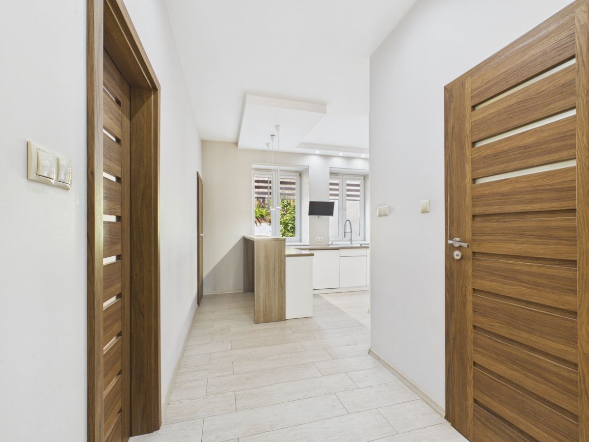 Bright hallway with light wood doors leading to a modern white kitchen with a window view outside far end