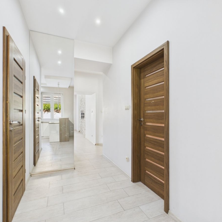 Bright hallway with white walls, light tile floor, and wood-paneled doors; a tall mirror reflects a kitchen area at the end.