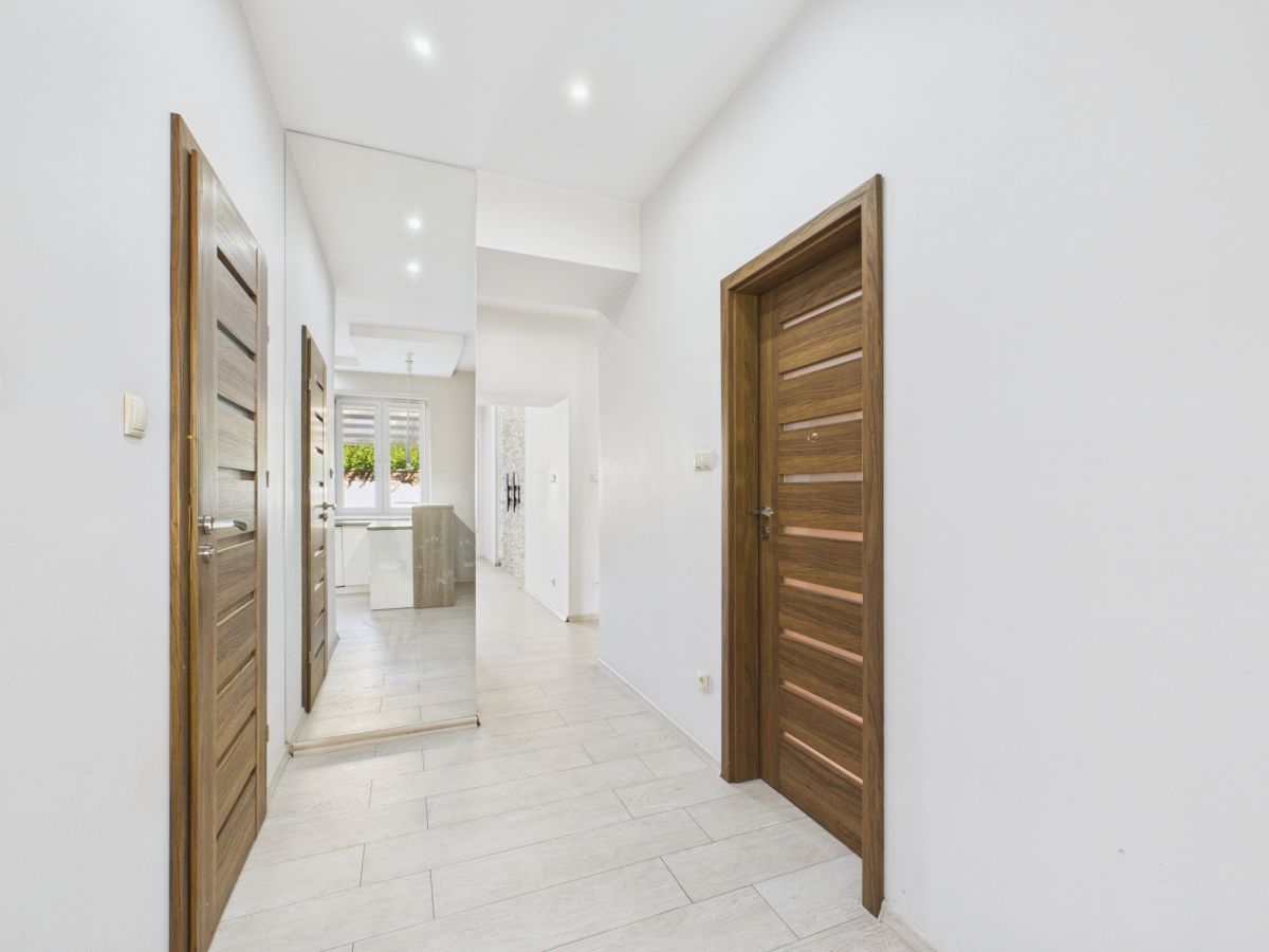 Bright hallway with white walls, light tile floor, and wood-paneled doors; a tall mirror reflects a kitchen area at the end.