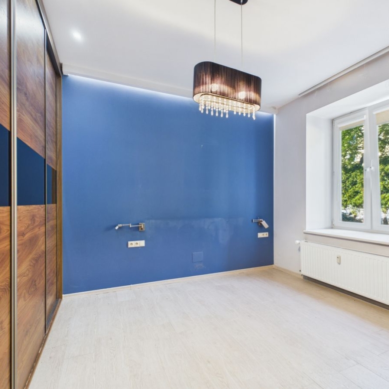 Empty bedroom with a bold blue feature wall, wooden wardrobe, and a hanging chandelier beside a large window in daylight.