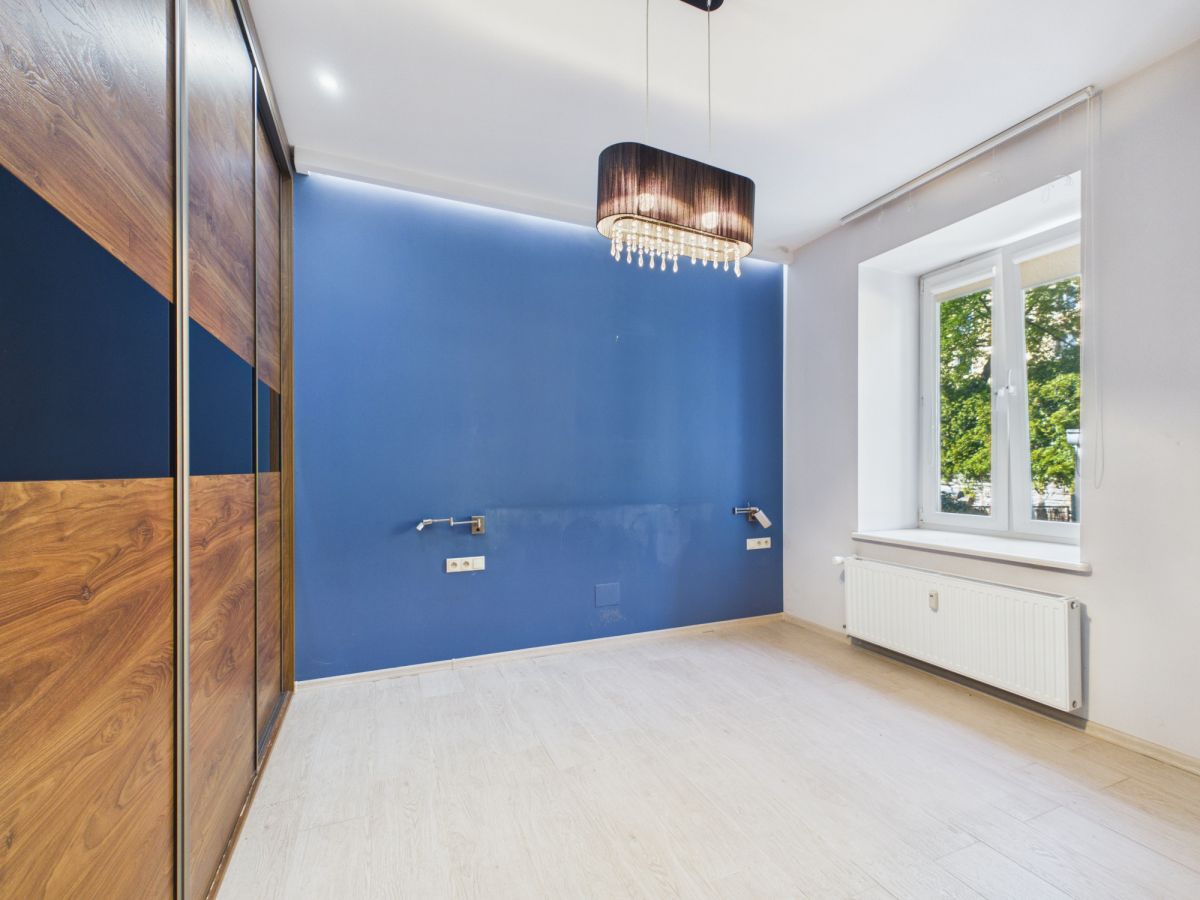 Empty bedroom with a bold blue feature wall, wooden wardrobe, and a hanging chandelier beside a large window in daylight.