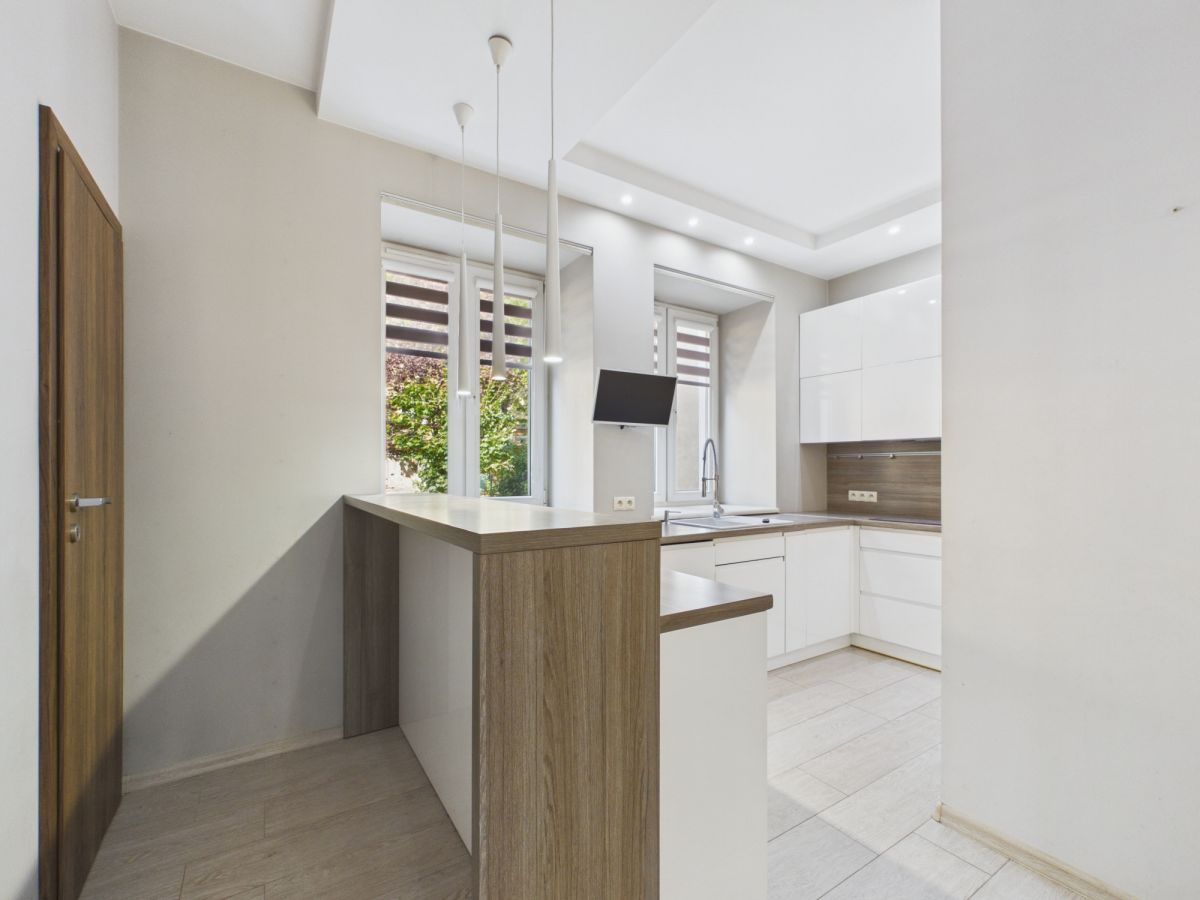 Bright modern kitchen with white cabinets and an oak-toned island, underpendant lights, and large windows overlooking greenery.