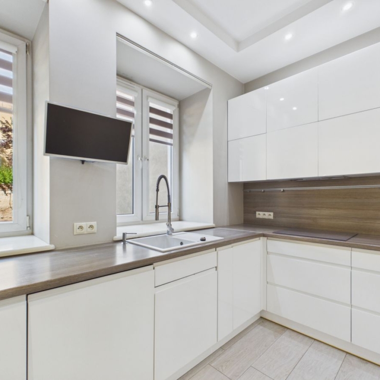 Modern white kitchen with wooden countertops, a double window over the sink, and a wall-mounted TV nearby.