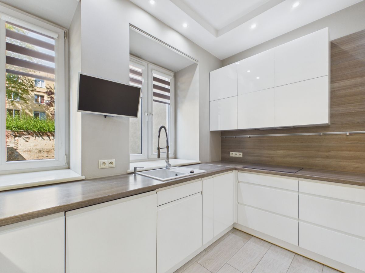 Modern white kitchen with wooden countertops, a double window over the sink, and a wall-mounted TV nearby.
