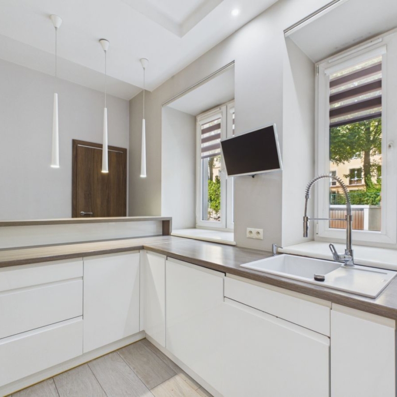 Modern L-shaped kitchen with white cabinets, wood countertop, pendant lights, and a wall-mounted TV by a large window.