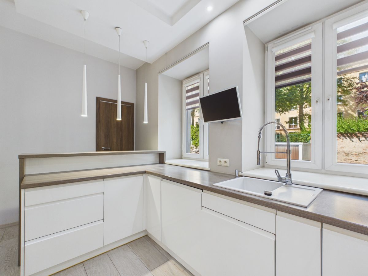 Modern L-shaped kitchen with white cabinets, wood countertop, pendant lights, and a wall-mounted TV by a large window.
