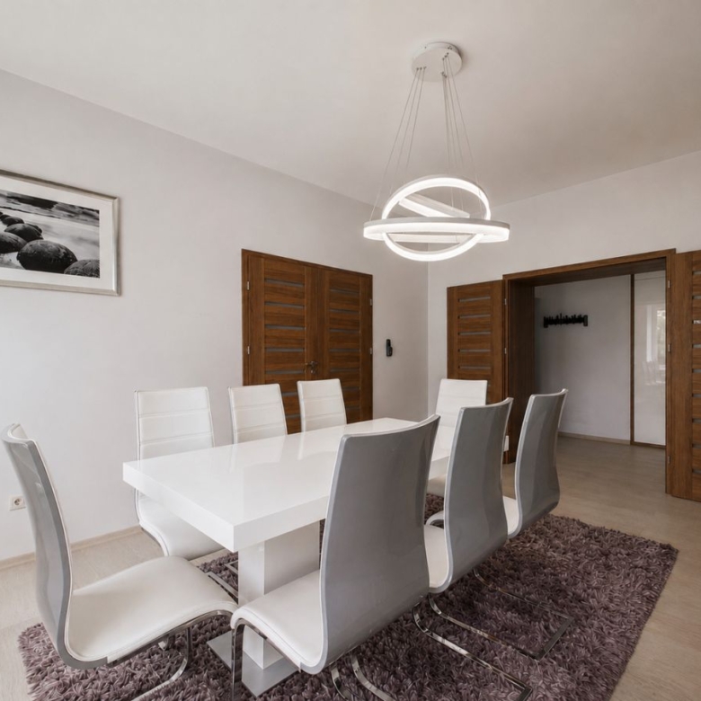 Modern dining room with a glossy white table, white and gray chairs, wooden double doors, and a circular ring chandelier above a purple rug.