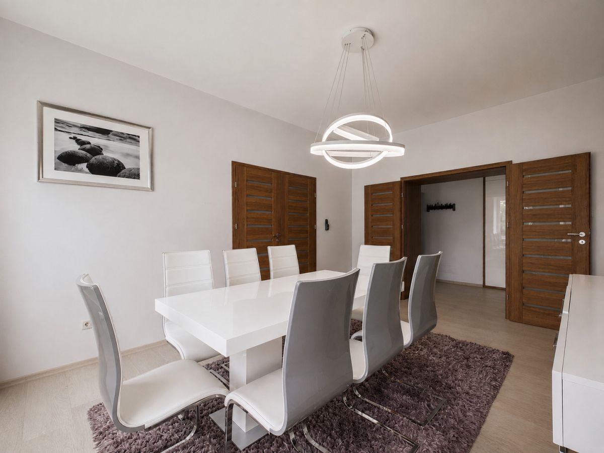Modern dining room with a glossy white table, white and gray chairs, wooden double doors, and a circular ring chandelier above a purple rug.