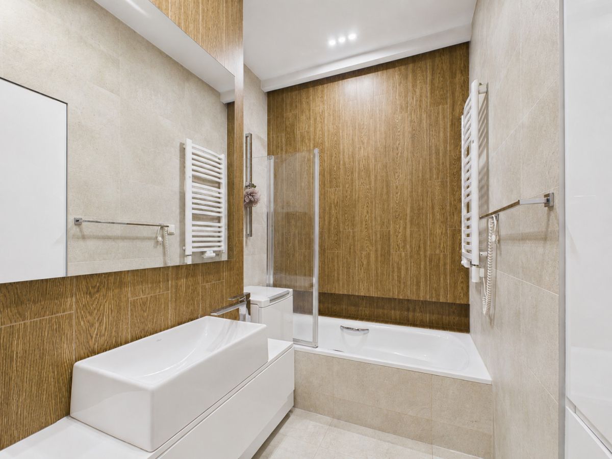 Modern white bathroom with a wood-paneled accent wall, wall-mounted sink, and bathtub with a glass shower screen.