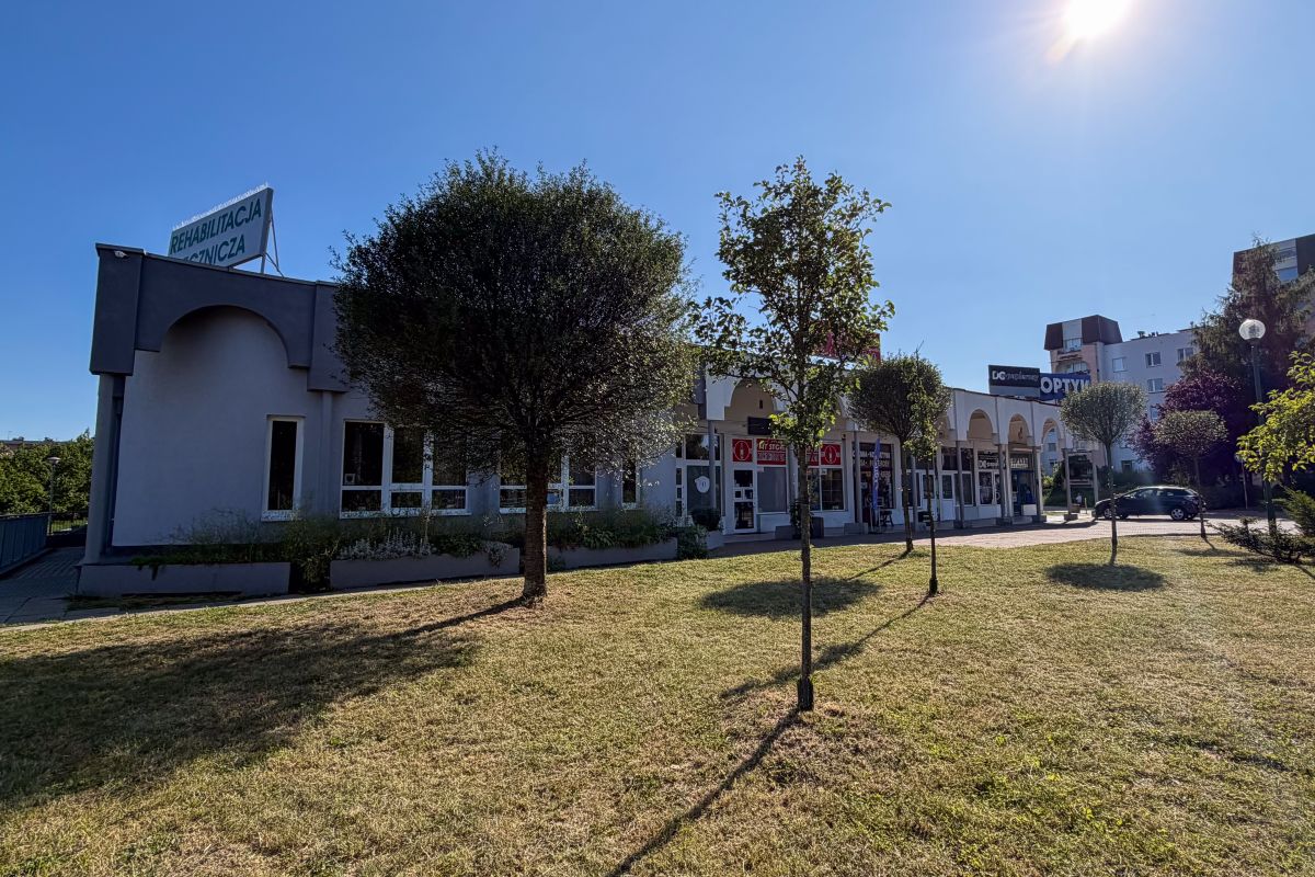 Row of storefronts with arches along a sunny plaza; a sign reads 'REHABILITACJA MEDYCZNA' atop the building, trees in the foreground.